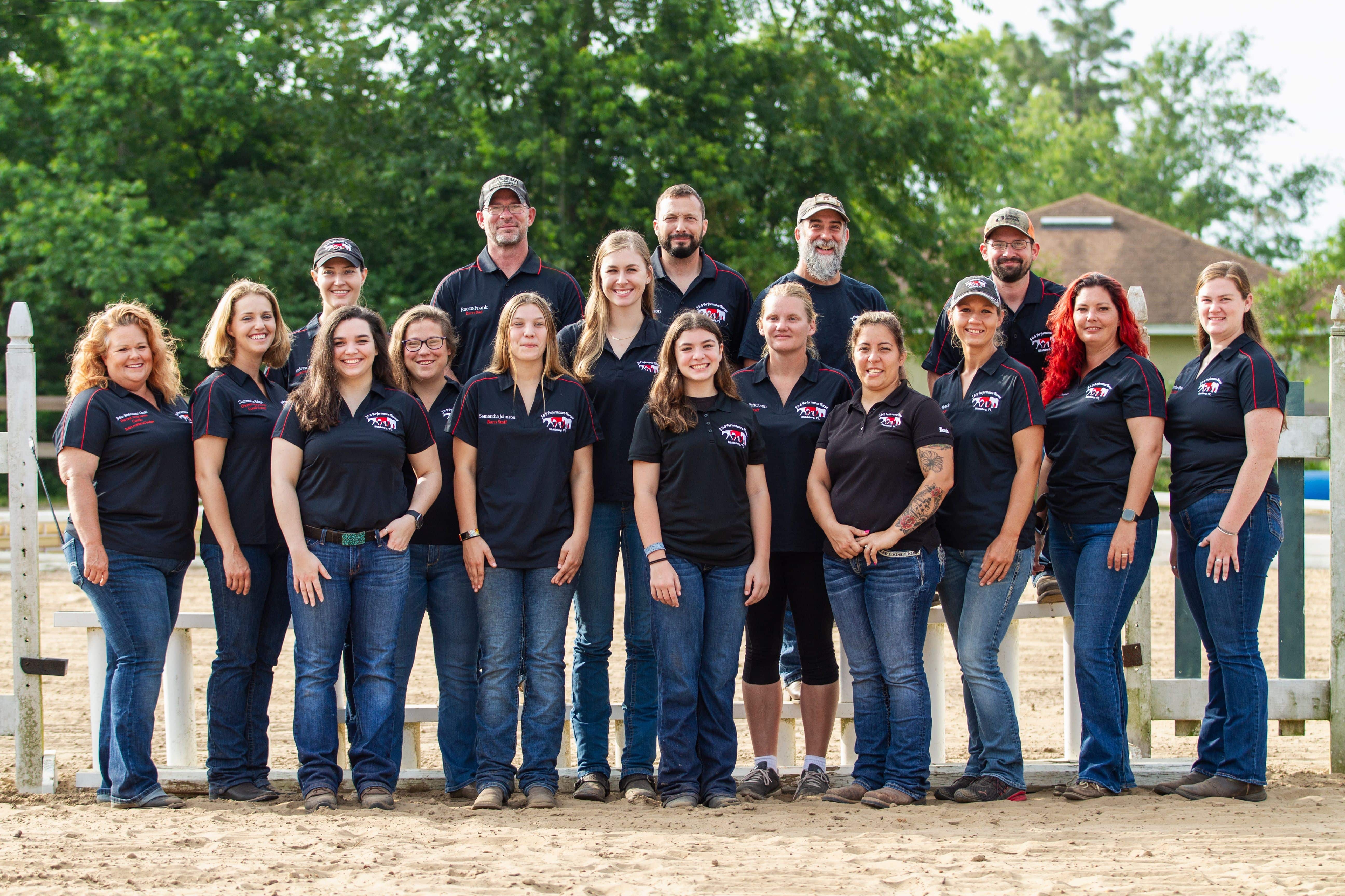 All of the staff and management at J & S are standing together for a group photo smiling. They are standing in front of a jump in the main arena.