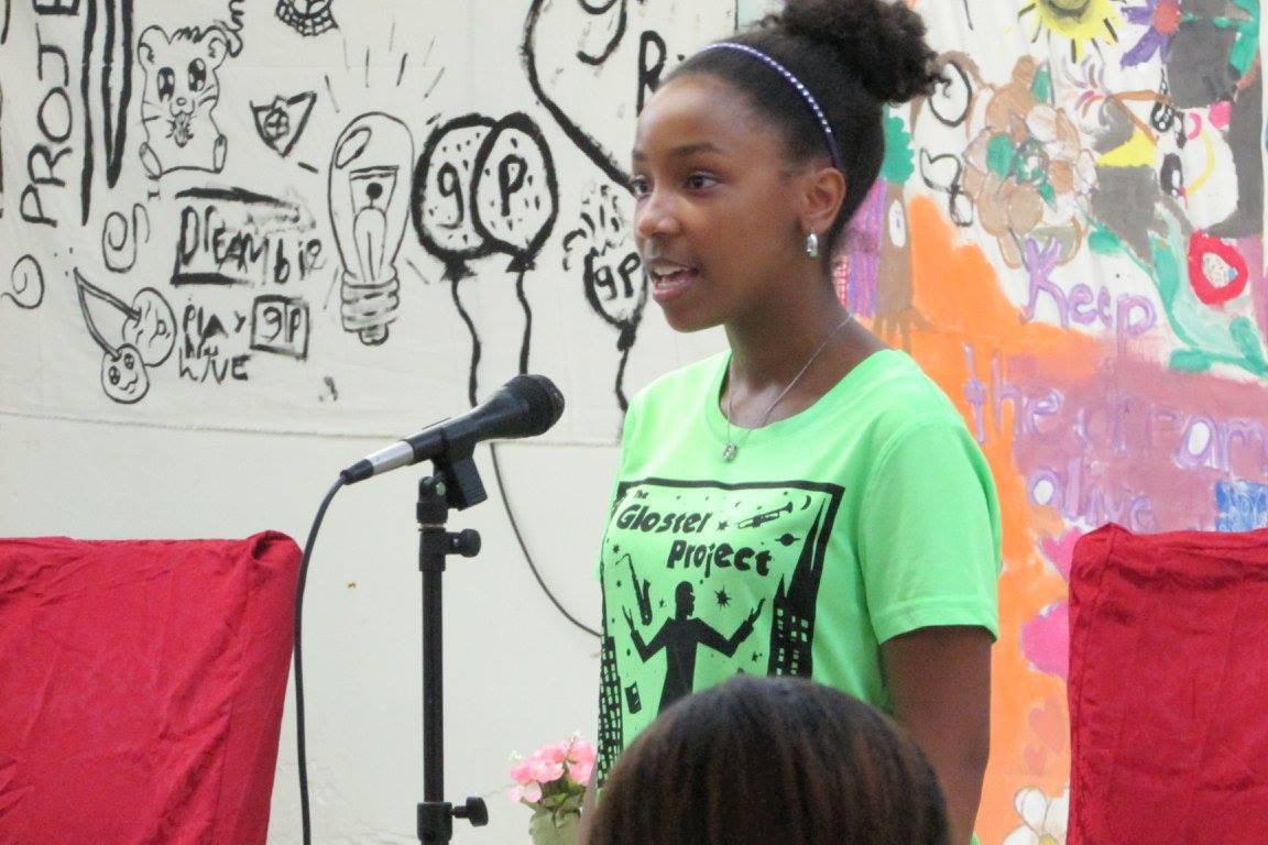 A girl stands at a microphone with drawings behind her ont he wall.