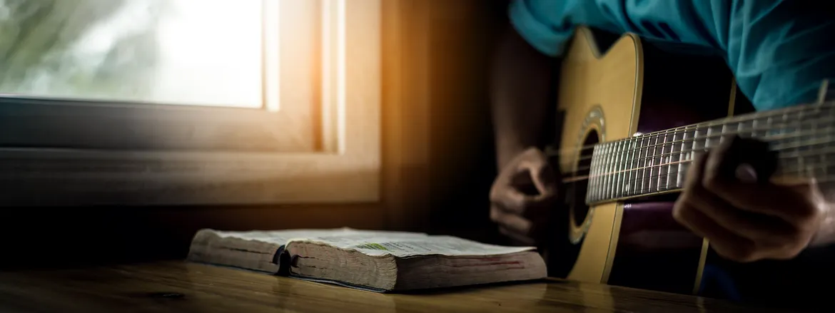 A person strums a guitar beside an open book on a wooden surface