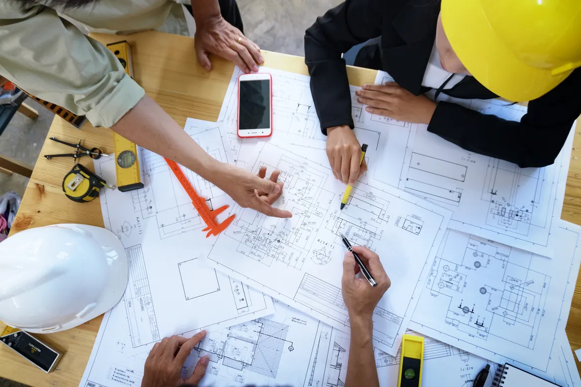 Professionals collaboratively review architectural blueprints on a table, using tools and a smartphone to discuss design details.