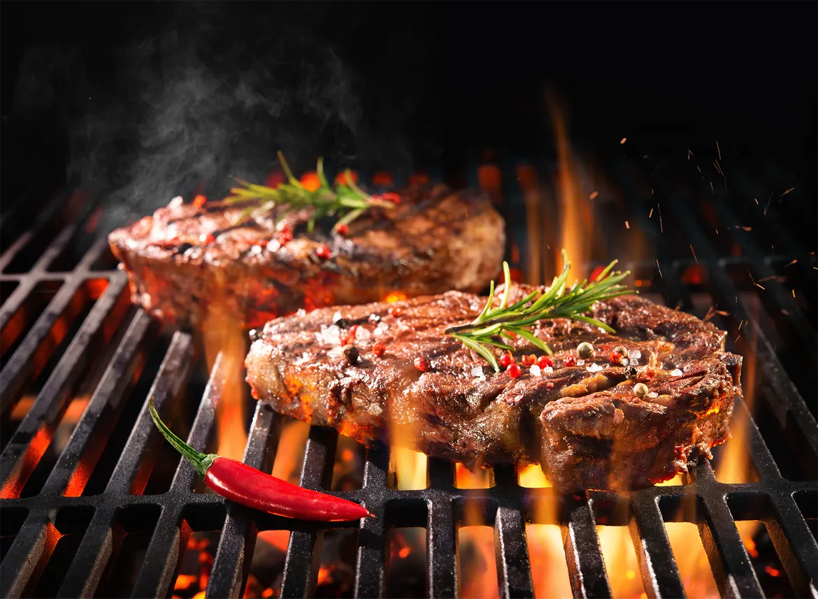 Grilled steaks searing over flames, garnished with herbs and spices